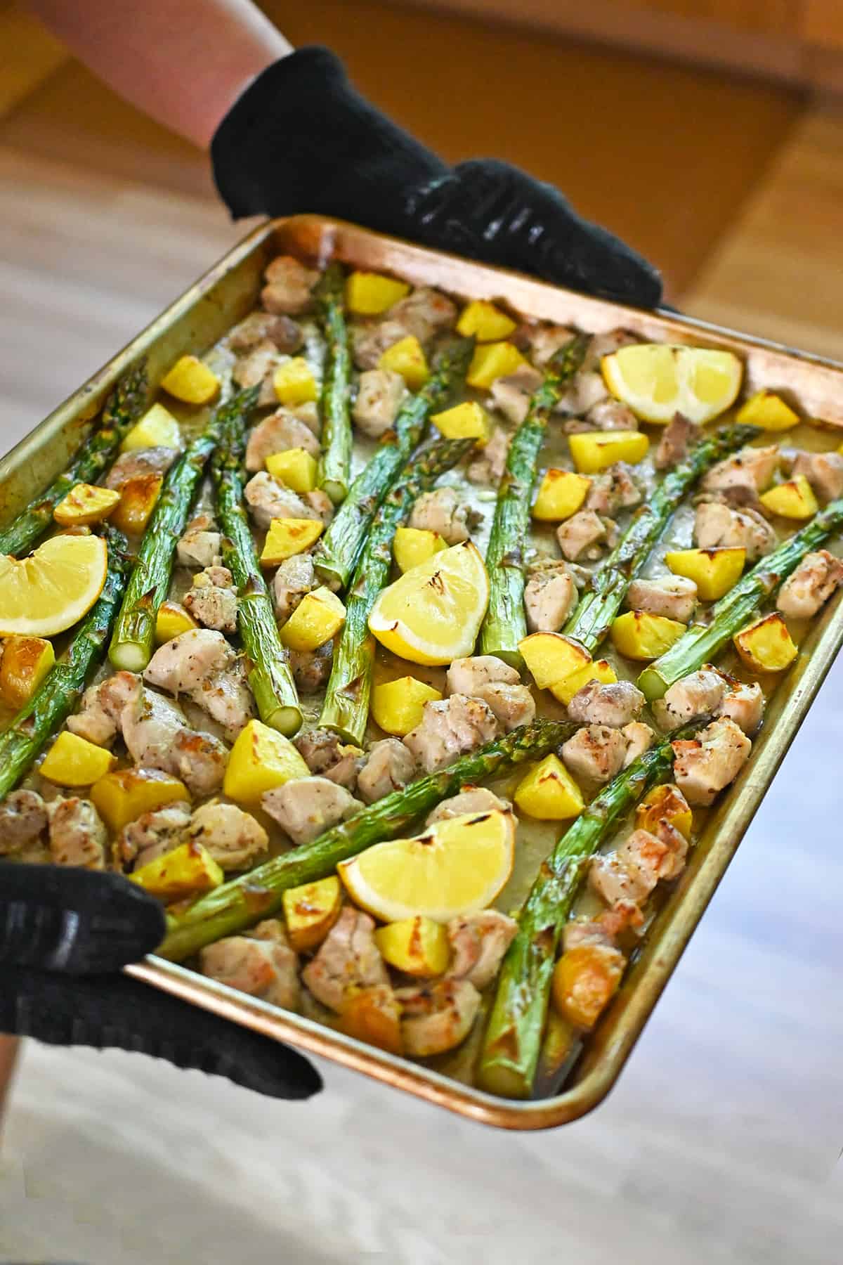Two hands in oven mitts are holding a sheet pan filled with golden brown chicken, potatoes, asparagus, and lemon wedges.