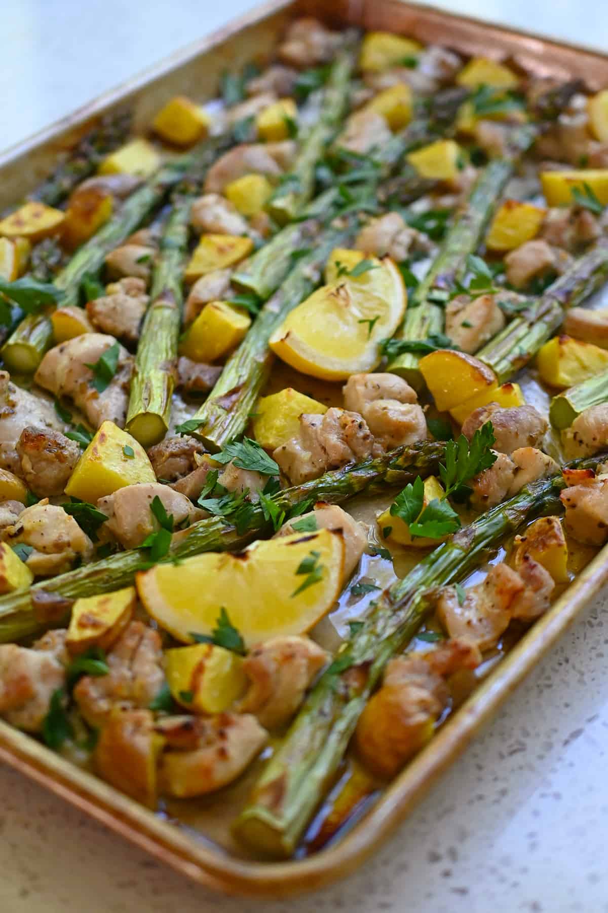 A side view of a Greek sheet pan chicken dinner with asparagus, potatoes, chicken, and lemon wedges.