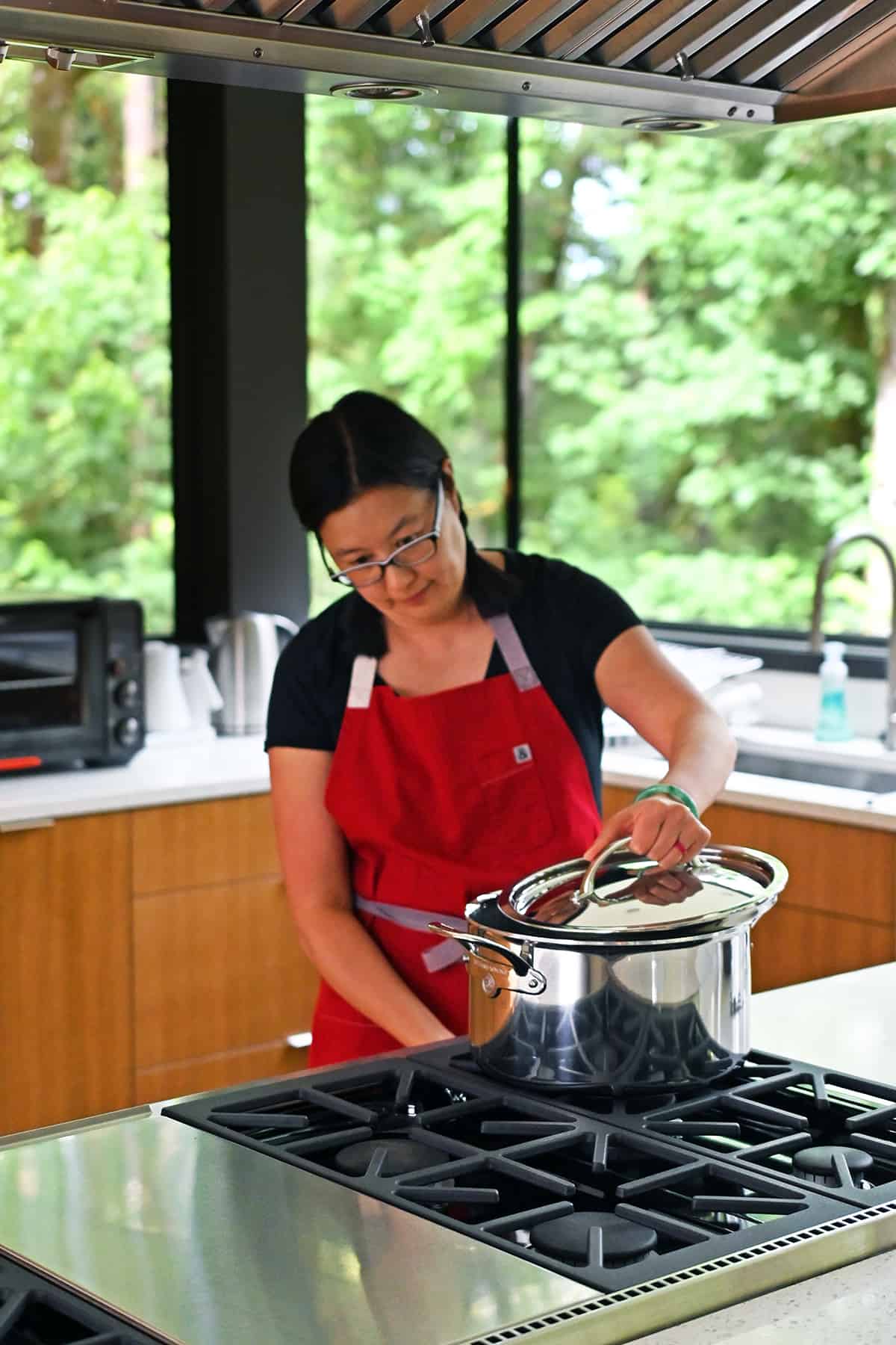 An Asian woman in a red apron is putting a lid on a big stockpot on a stovetop.