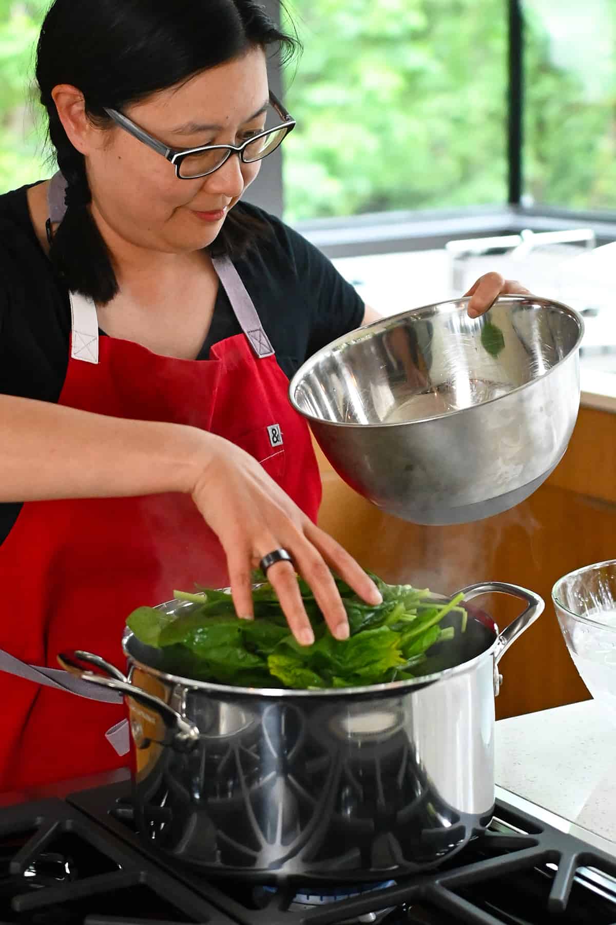 An Asian woman in a red apron is adding raw spinach to a large pot of boiling water.