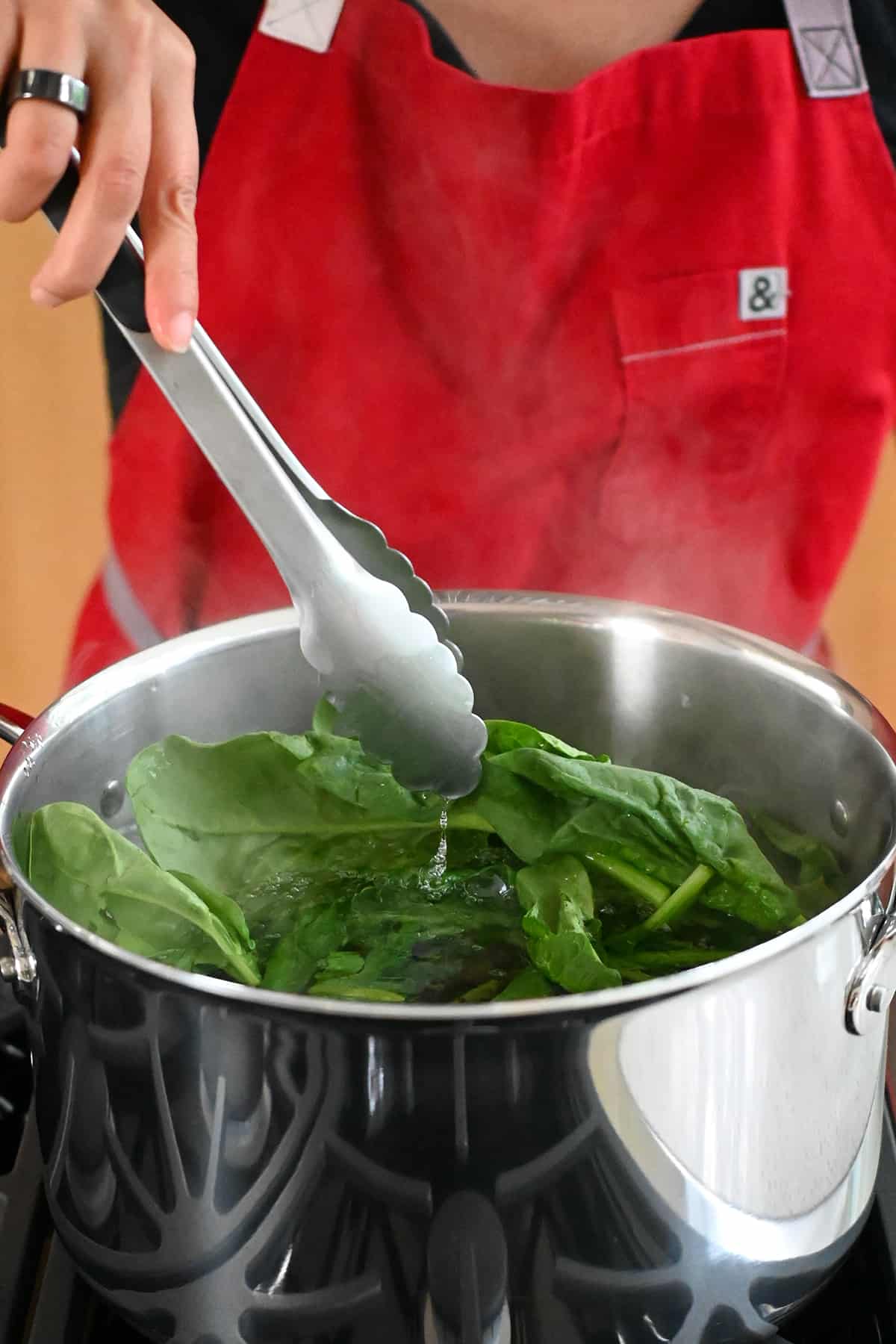 A pair of tongs is adding raw spinach to a large pot of boiling water to make a Korean spinach side dish.