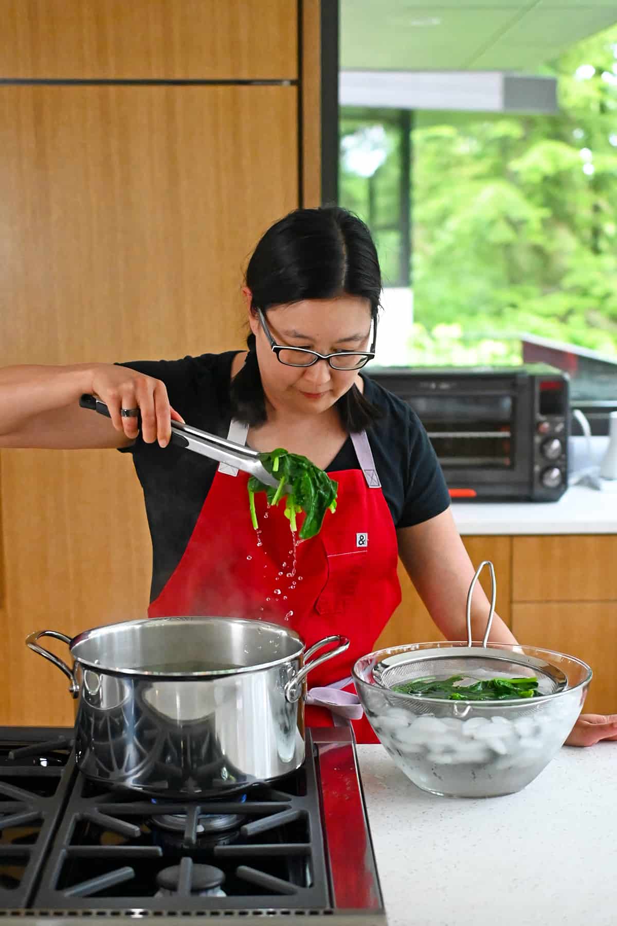 An Asian woman is transferring cooked spinach from a pot of boiling water to a glass bowl filled with ice water and a strainer.