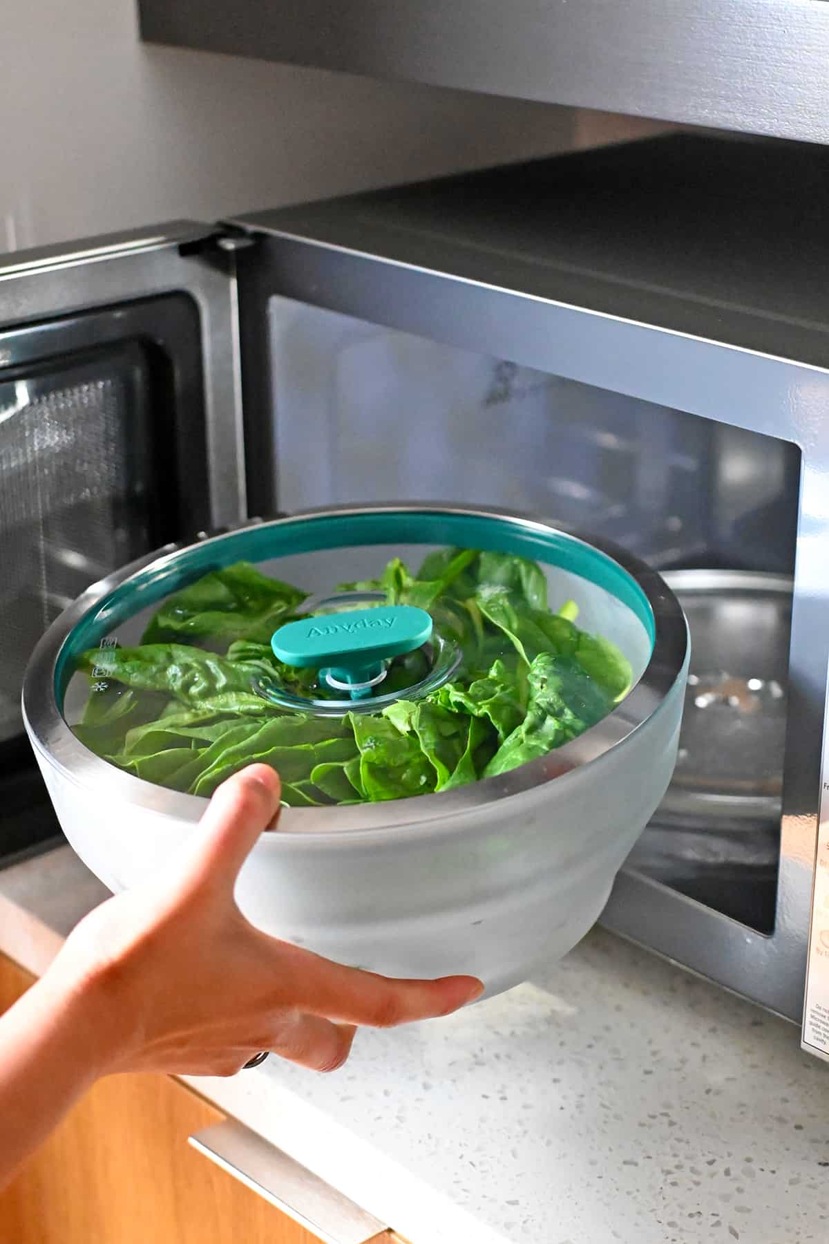 A hand is placing an extra large glass Anyday microwave container filled with raw spinach into an open microwave.