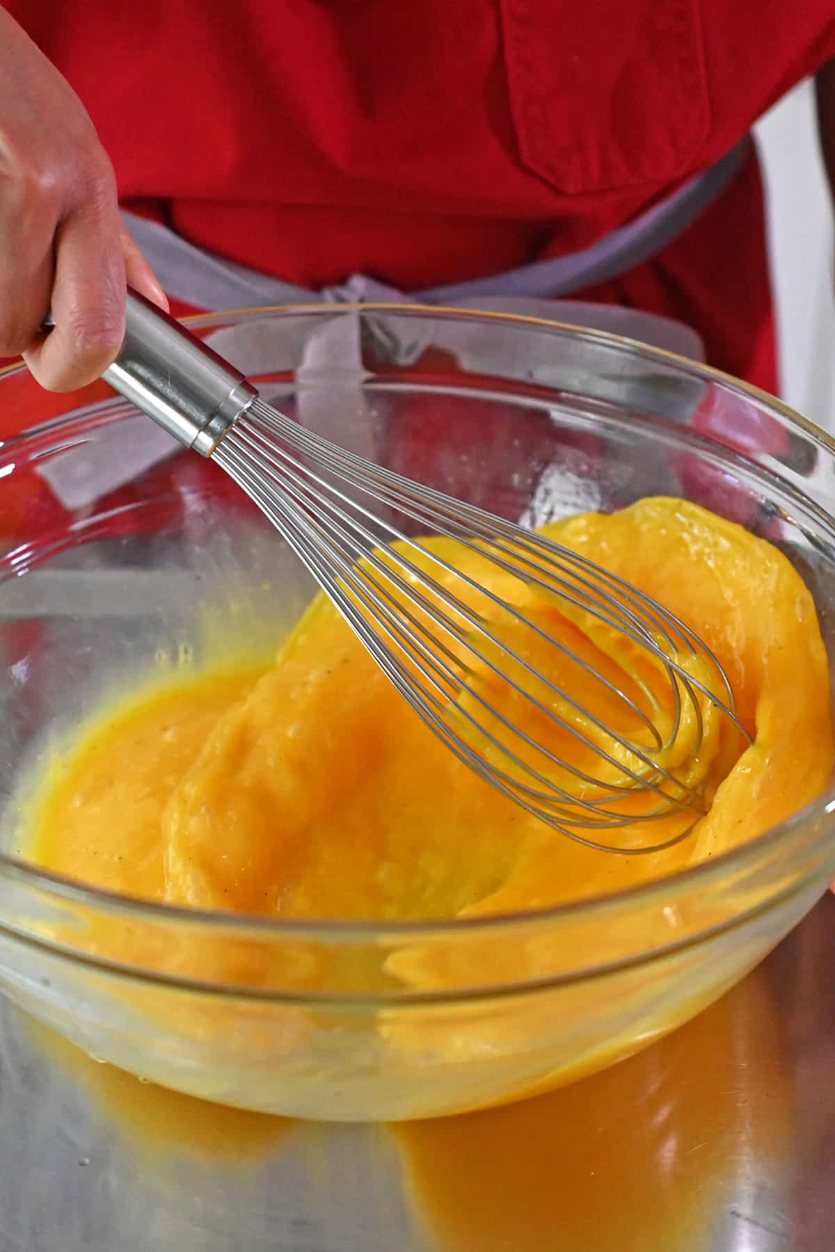 A closeup of whisking raw eggs in a large glass mixing bowl with a metal whisk.