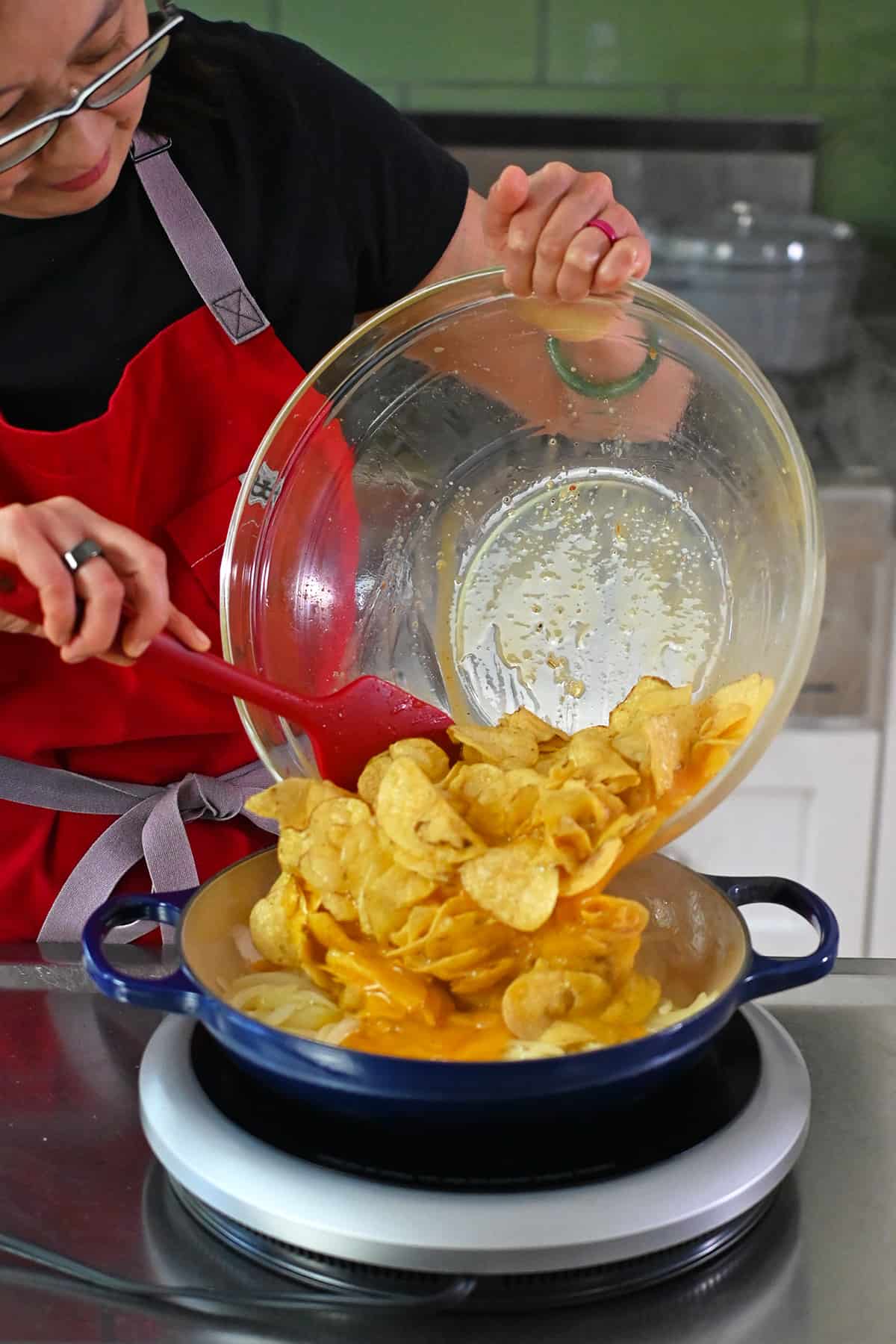 An Asian woman in glasses is adding a bowl of whisked eggs and potato chips to a blue skillet with sautéed onions to make a quick Spanish tortilla.