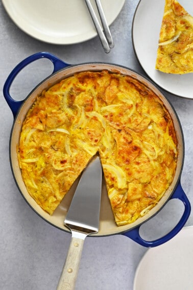 An overhead shot of a skillet filled with a Spanish tortilla made with potato chips with a missing slice that is on a plate next to the pan.
