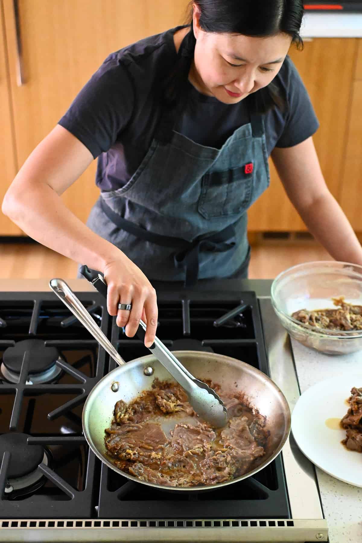 An Asian woman is cooking paleo bulgogi in a skillet on the stovetop in batches.