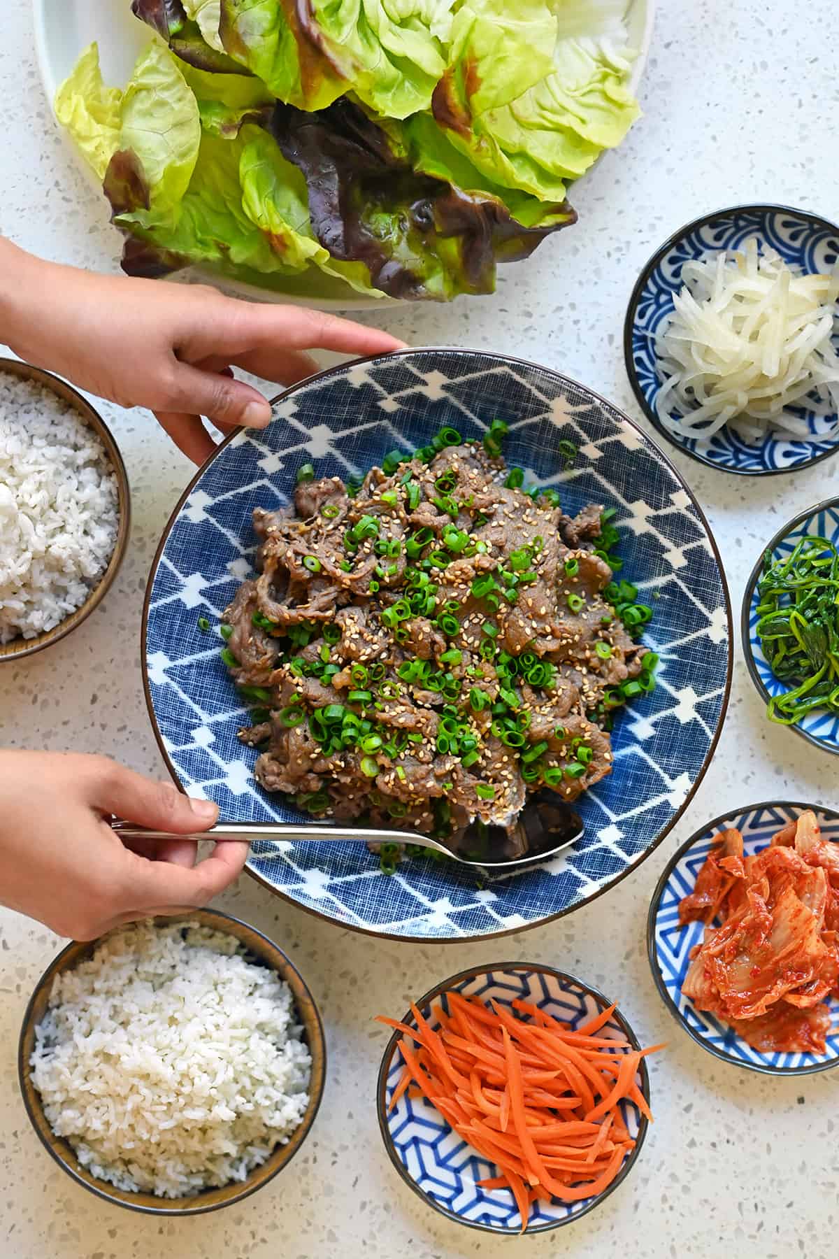 A hand is placing a serving spoon into a bowl of paleo bulgogi. There are small bowls of vegetables and steamed rice surrounding the beef.