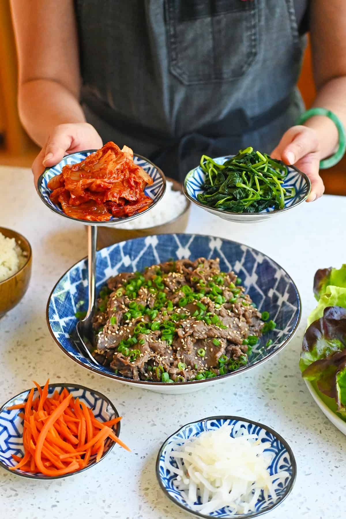 Two hands holding small bowls of kimchi and cooked spinach above a bowl of bulgogi and other small plates of banchan.