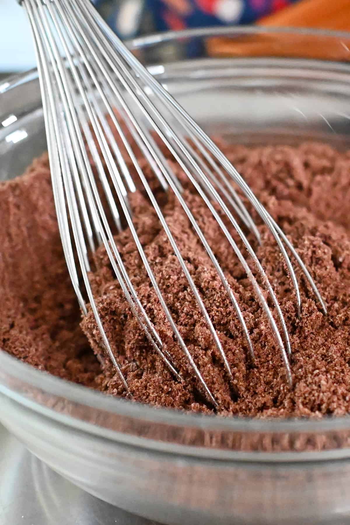A metal whisk is stirring the dry ingredients in a bowl for chocolate zucchini muffins.