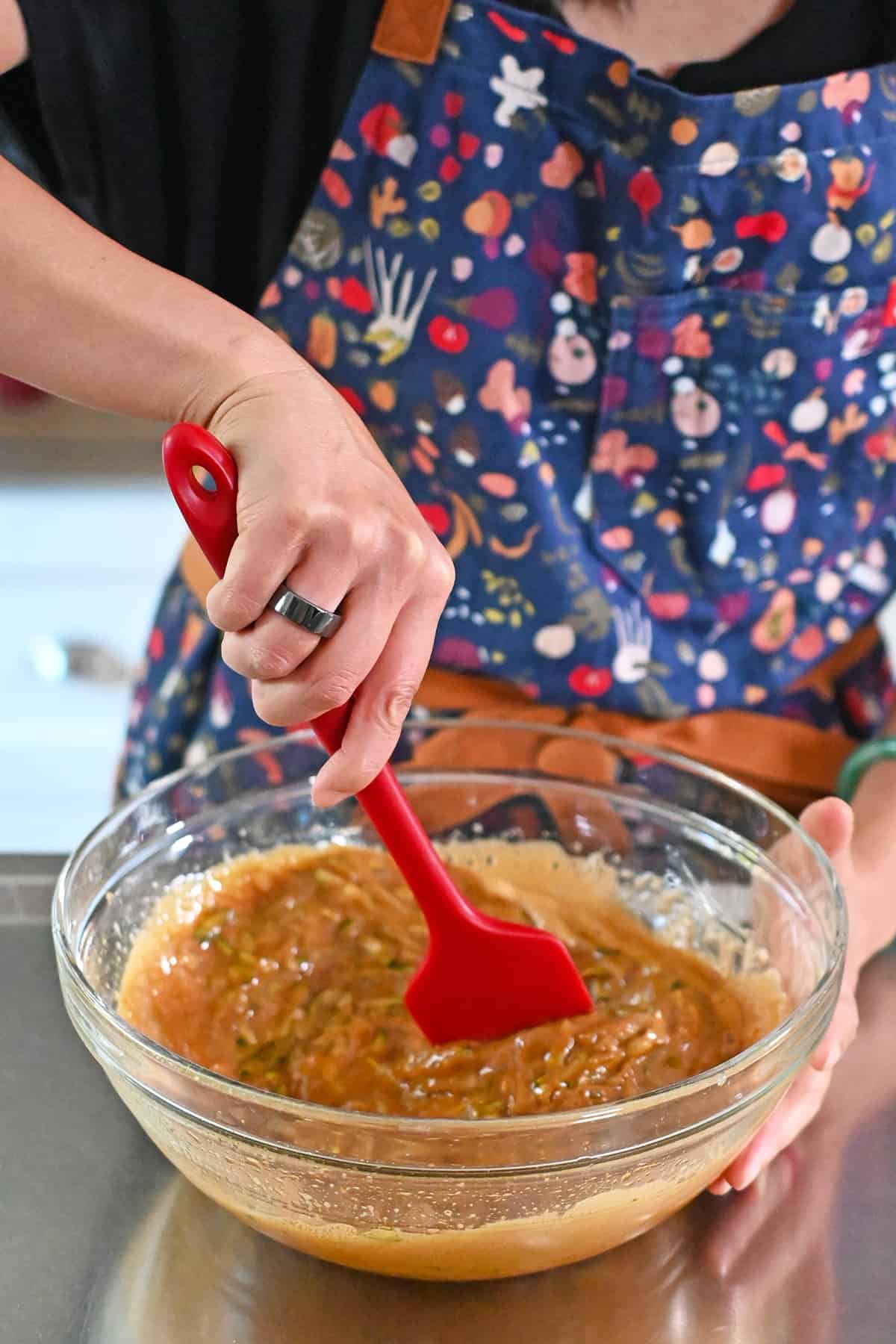 Stirring the shredded zucchini into the coconut sugar and egg mixture with a red silicone spatula.