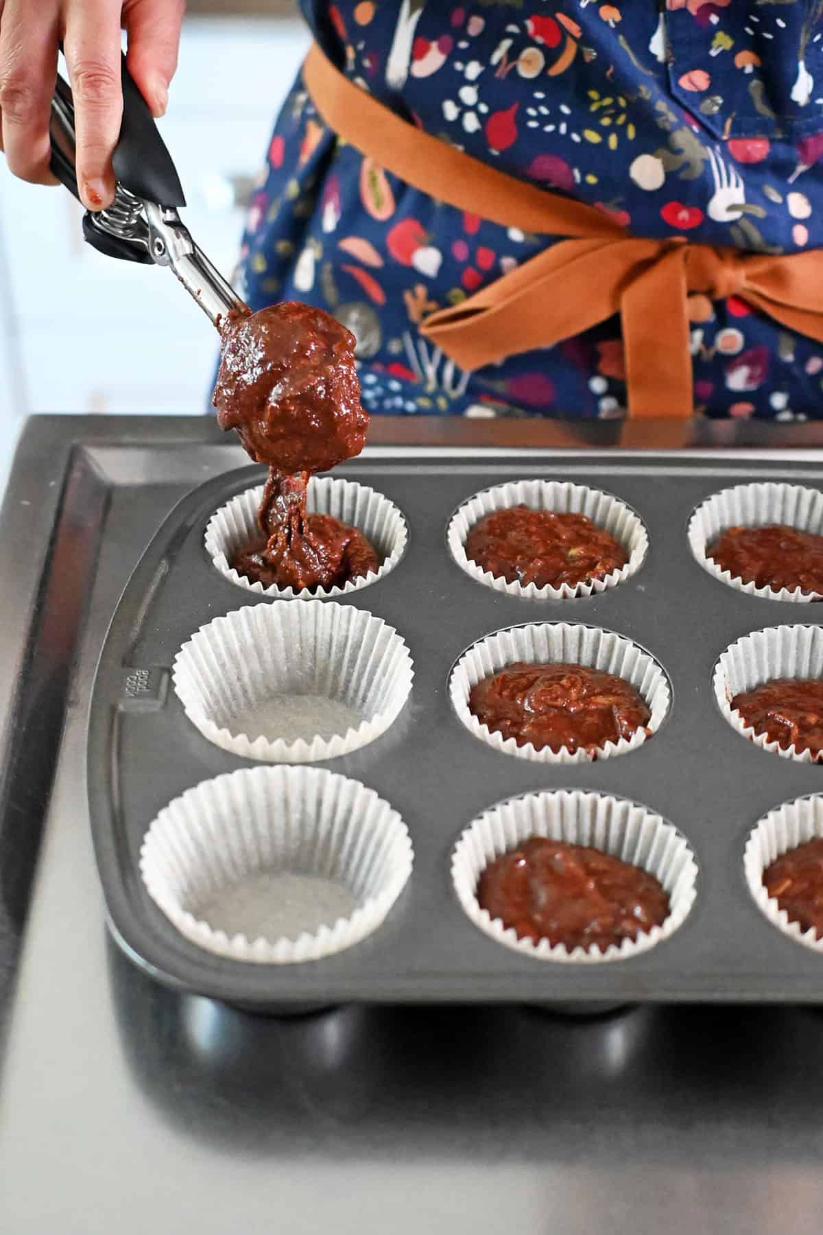 Using an ice cream scooper to transfer the chocolate zucchini muffin batter into a parchment paper lined muffin tin.