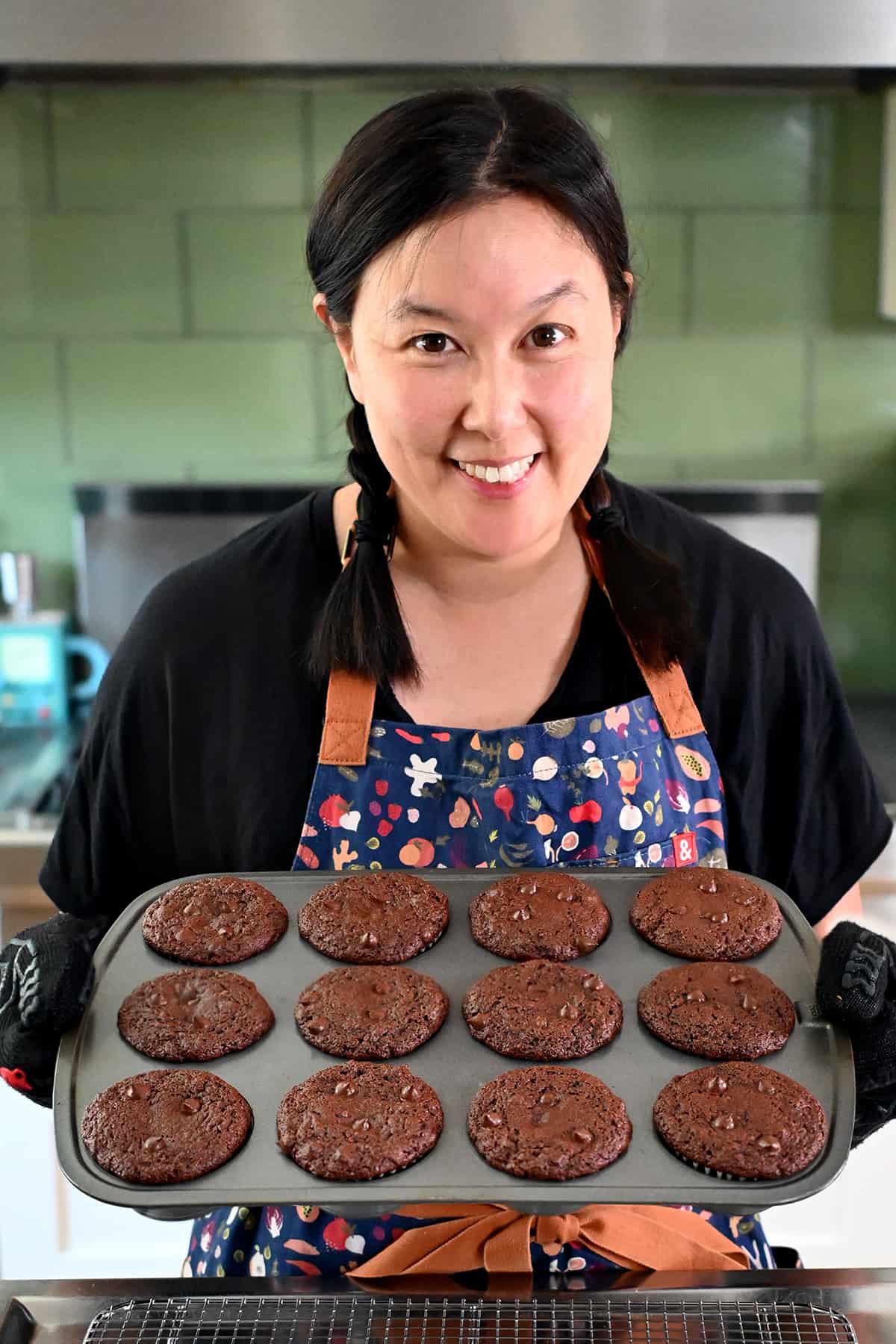 A smiling Asian woman in braids is holding up a muffin pan filled with paleo chocolate zucchini muffins.
