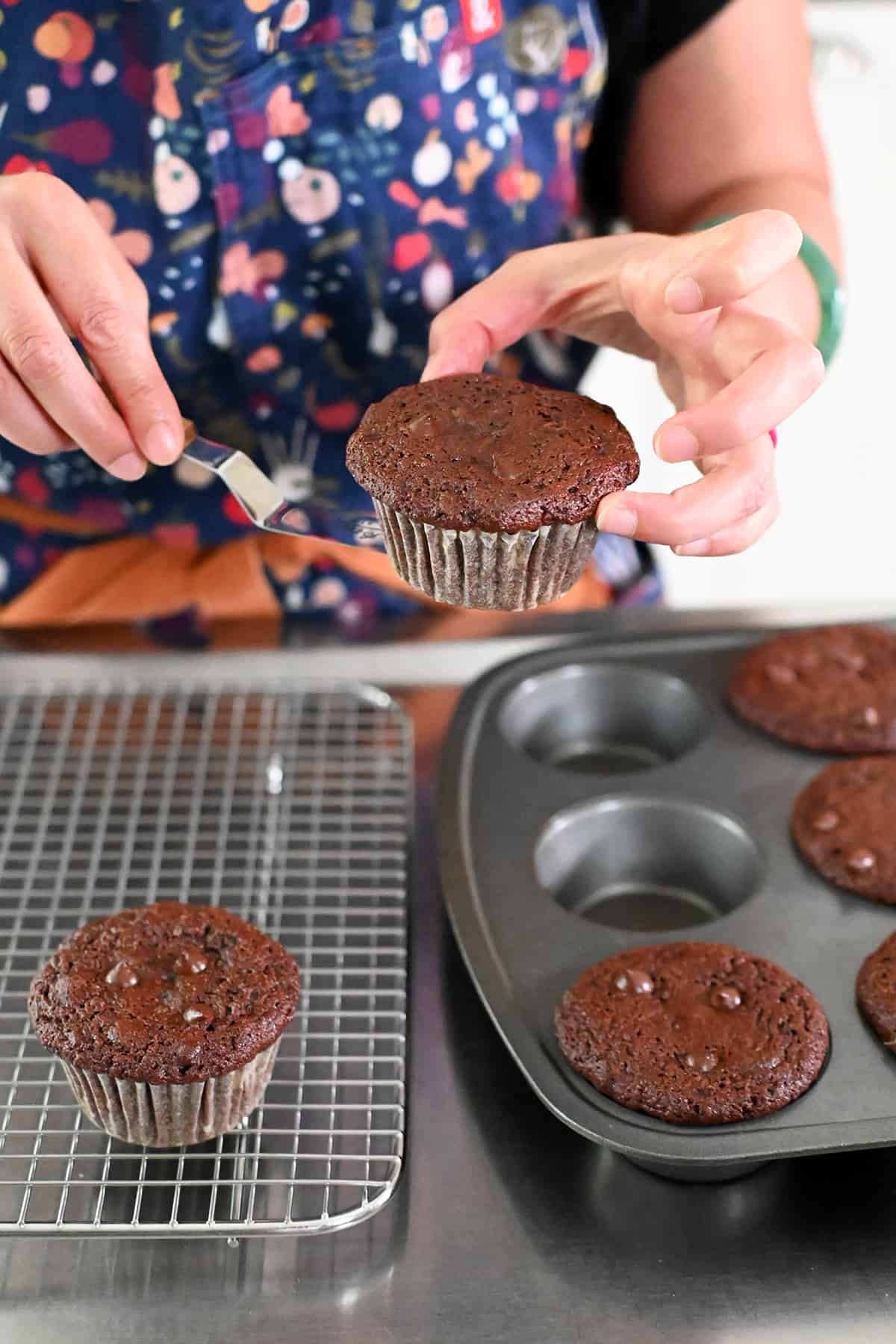Lifting a freshly baked chocolate zucchini muffin from the tin and putting it on a cooling rack.