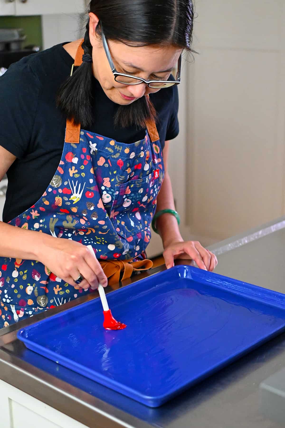 A smiling Asian woman is brushing oil on a blue rimmed baking sheet to make sheet pan eggs.