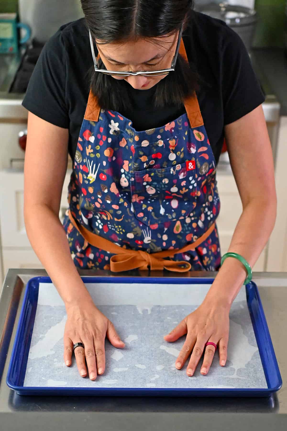 An Asian woman is placing a piece of parchment paper into a greased blue rimmed sheet pan,