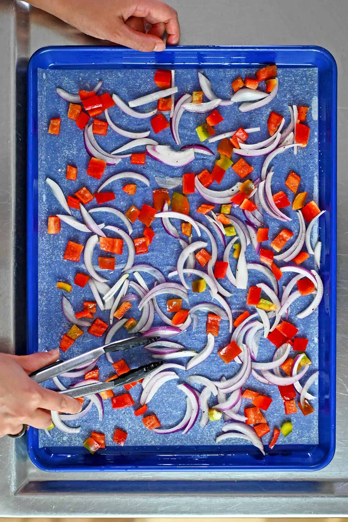 A pair of tongs is rearranging thinly sliced red onions and diced bell peppers in a single layer on a parchment lined rimmed baking sheet.