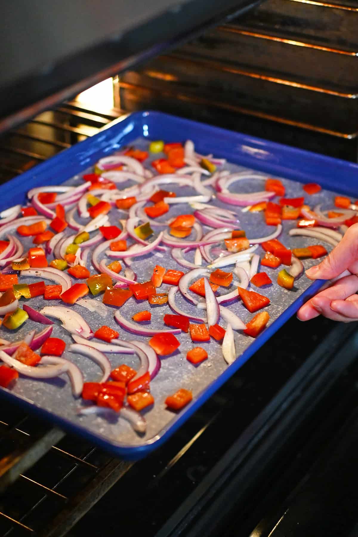 Putting a rimmed sheet pan with thinly sliced red onions and diced bell peppers into an open oven.