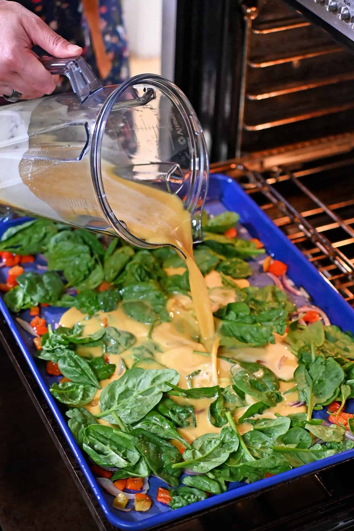 Pouring blended raw eggs from a Vitamix blender cup into a blue rimmed sheet pan filled with baby spinach, roasted sliced red onions, and roasted red bell peppers.