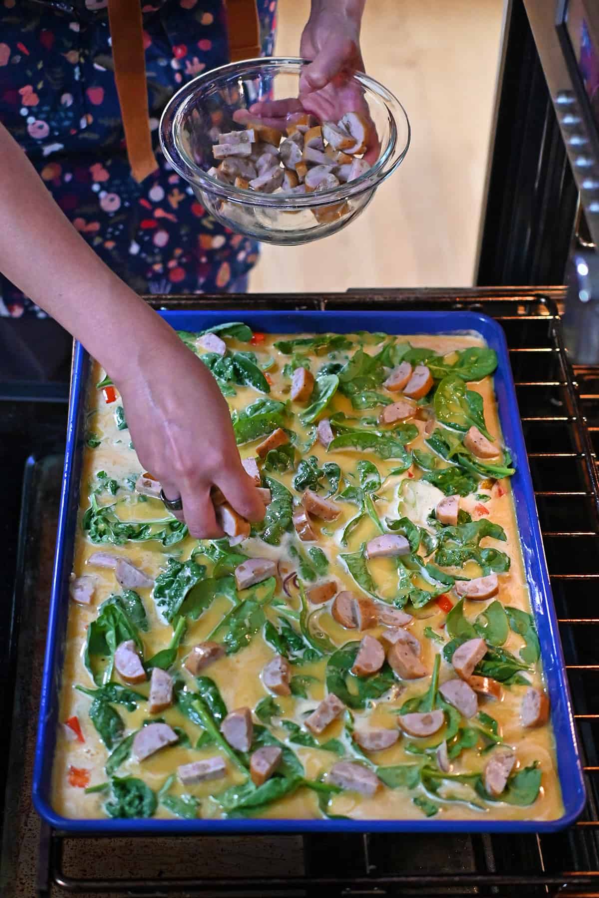 A hand is placing thinly sliced chicken apple sausage onto an unbaked blue sheet pan filled with raw scrambled eggs and vegetables in an open oven.