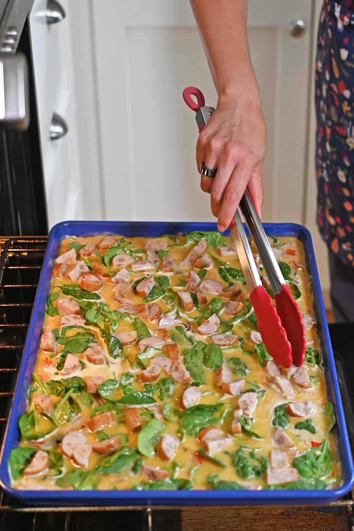 A pair of tongs is rearranging the ingredients on sheet pan eggs before it is put back in the oven.
