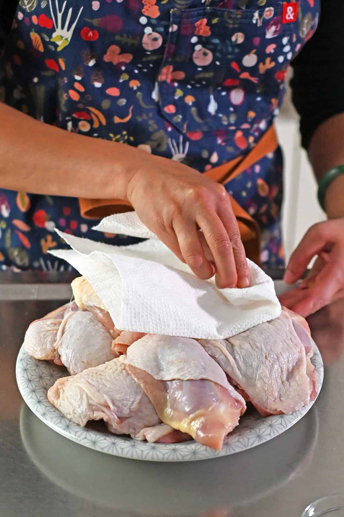 A person in a blue apron is blotting raw skin-on bone-in chicken thighs dry with a paper towel.