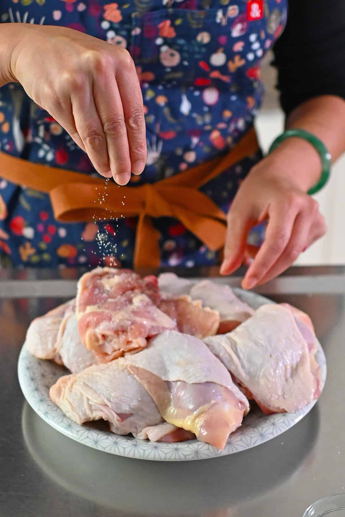 A person in a blue apron is sprinkling kosher salt on a plate filled with raw skin-on bone-in chicken thighs.