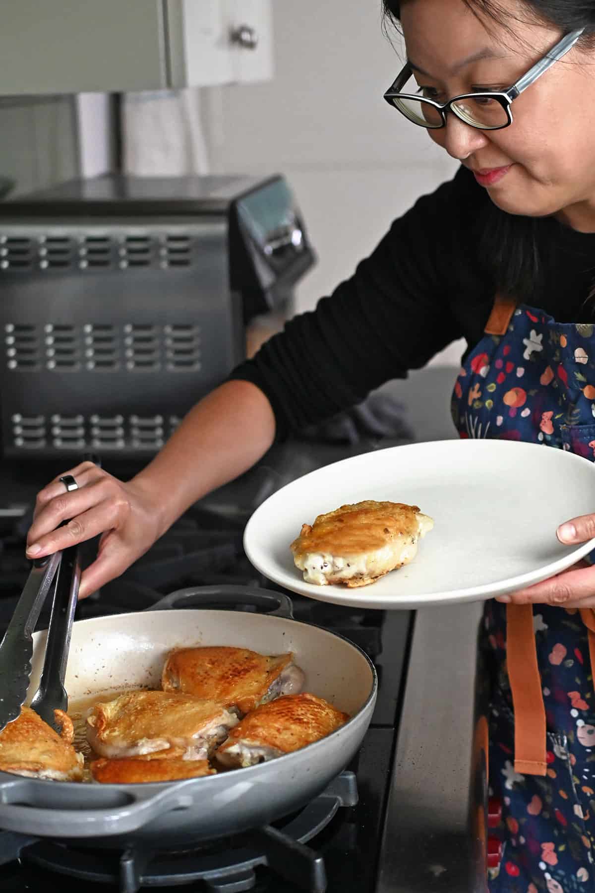 An Asian woman in glasses is transferring chicken thighs with golden brown skin from a skillet onto a plate with a pair of tongs.