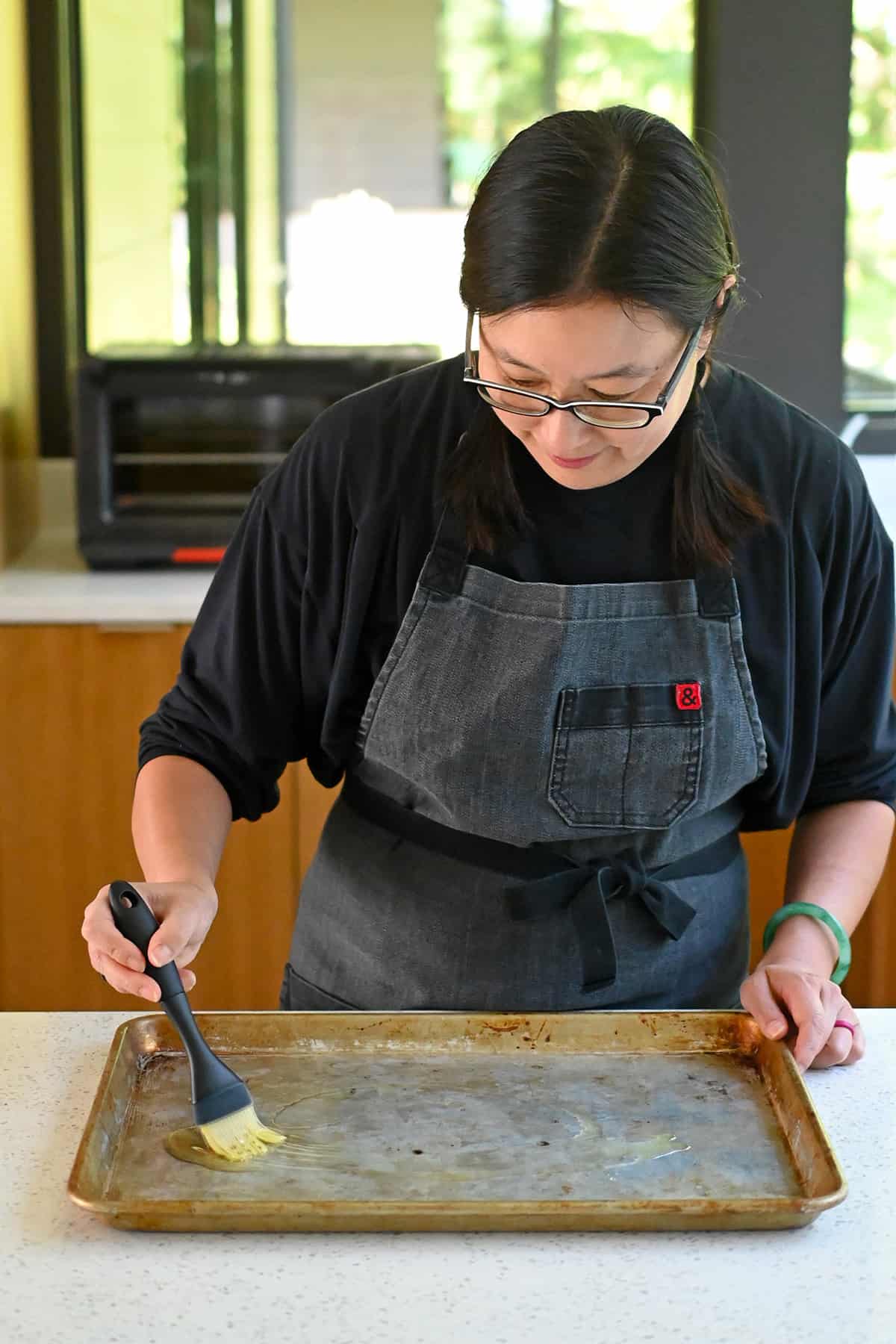 An Asian woman in glasses is brushing avocado oil on a rimmed baking sheet to grease it before making sheet pan cashew chicken.