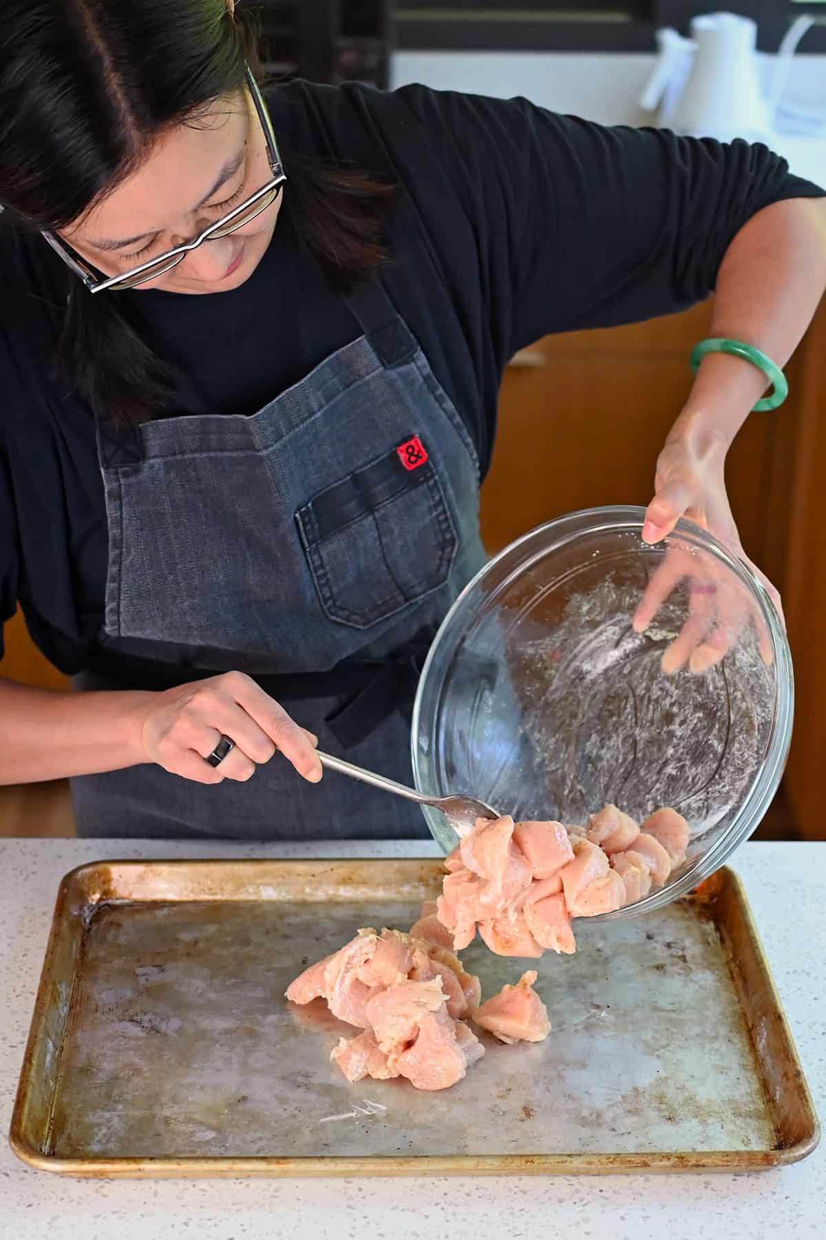 An Asian woman is adding seasoned raw chicken breast cubes to a greased rimmed baking sheet.
