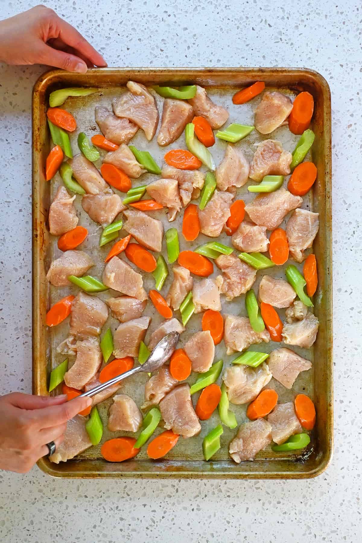 A spoon is rearranging raw chicken breast pieces, sliced carrots, and sliced celery in a single layer in a rimmed sheet pan.