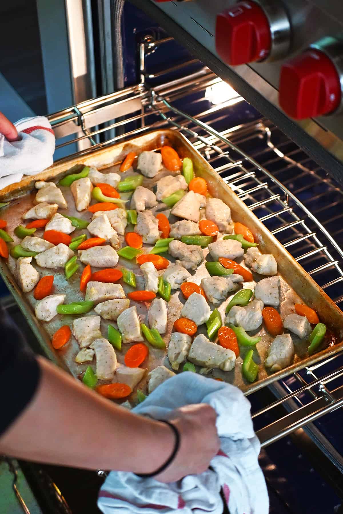 Removing a sheet pan from the oven with cooked chicken breast cubes, sliced carrots, and sliced celery.