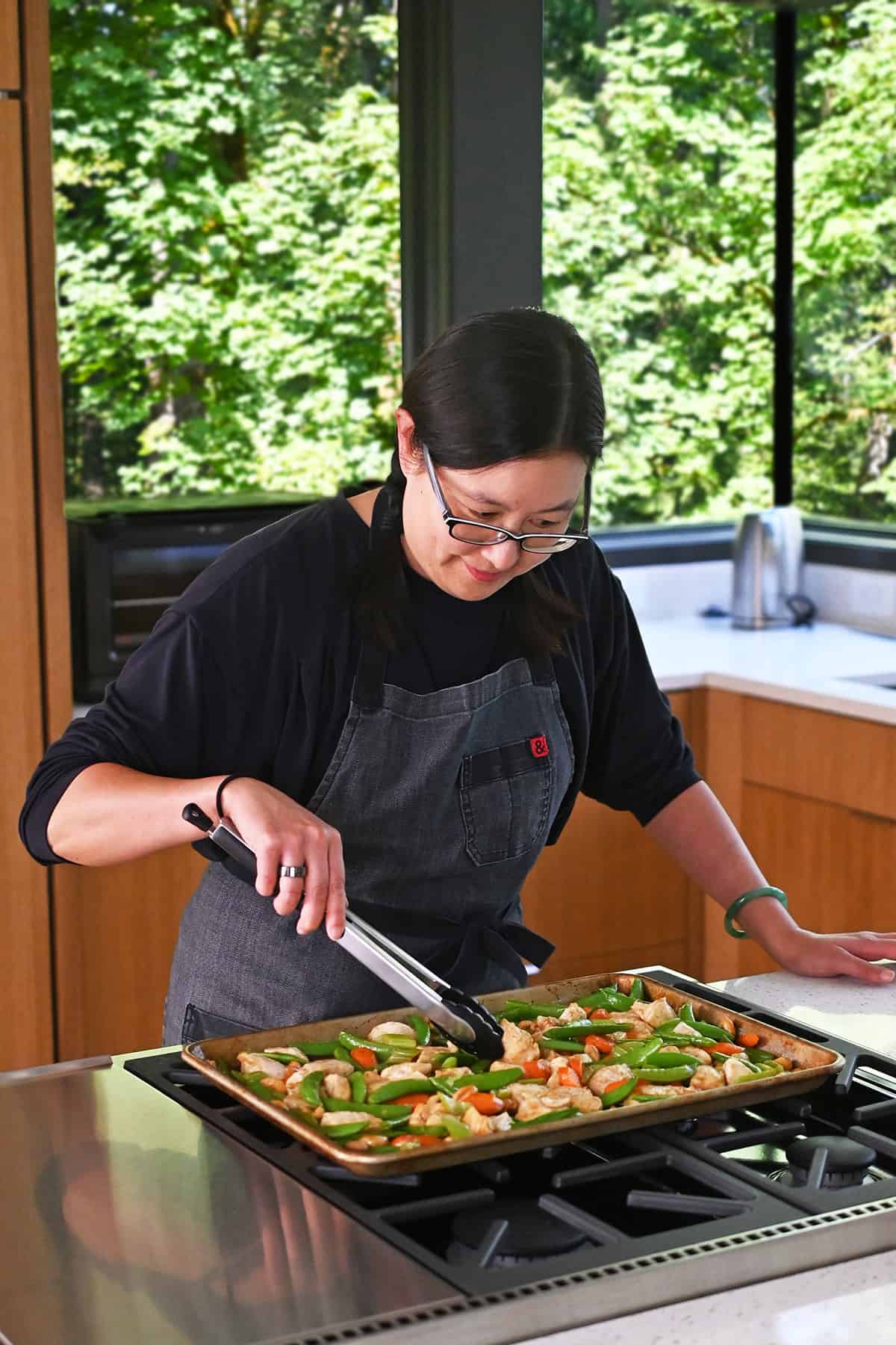 An Asian woman is using a pair of tongs to toss the sheet pan cashew chicken before serving.
