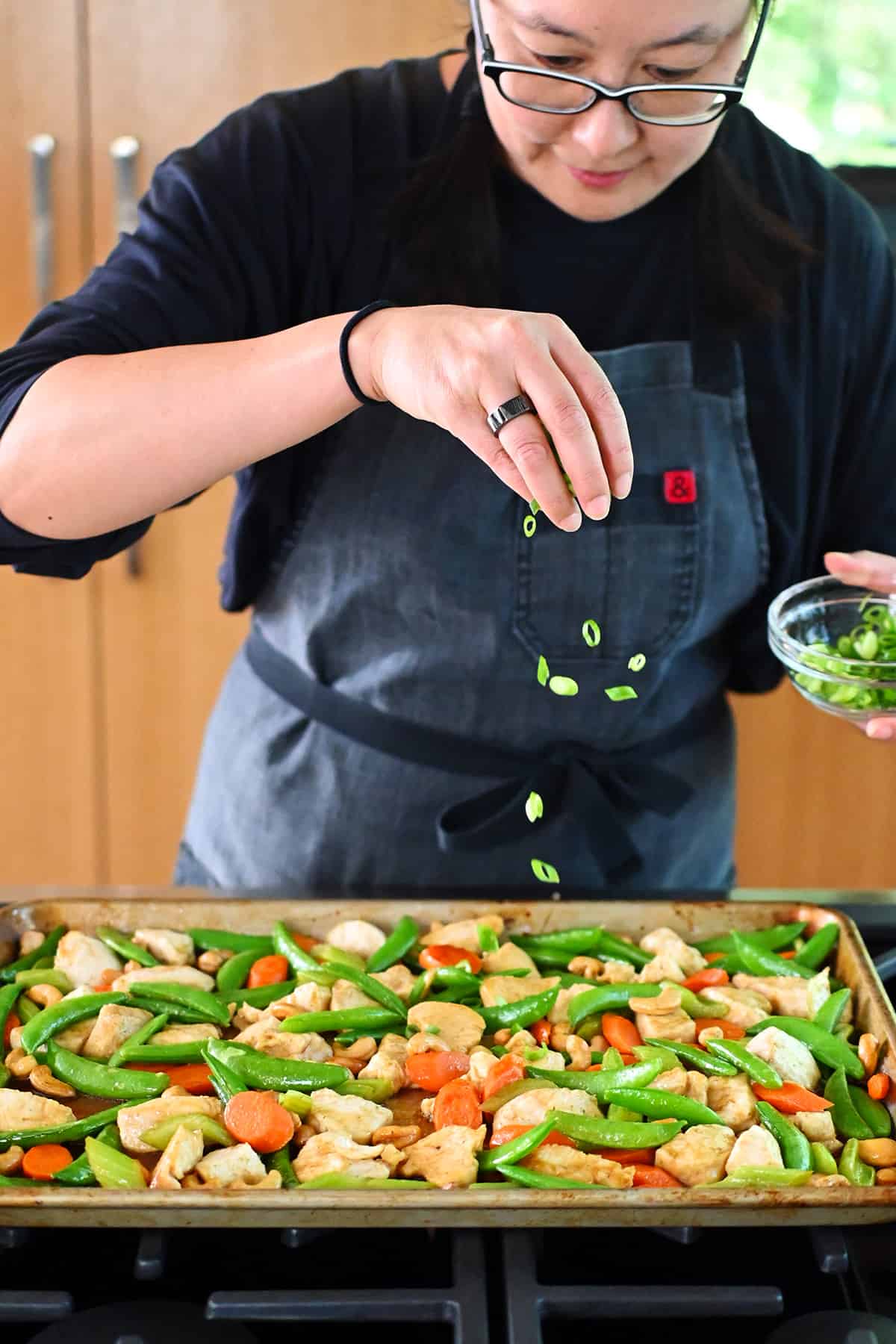 An Asian woman is sprinkling sliced green onions on top of a tray of Sheet Pan Cashew Chicken.