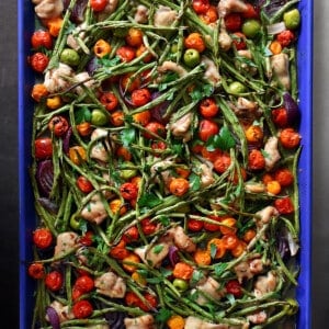 An overhead shot of a blue sheet pan filled with cooked cubed chicken thighs, roasted green beans, roasted cherry tomatoes, red onion wedges, green olives, fresh herbs, and a drizzle of balsamic vinegar.