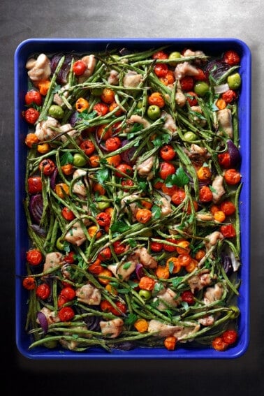 An overhead shot of a blue sheet pan filled with cooked cubed chicken thighs, roasted green beans, roasted cherry tomatoes, red onion wedges, green olives, fresh herbs, and a drizzle of balsamic vinegar.