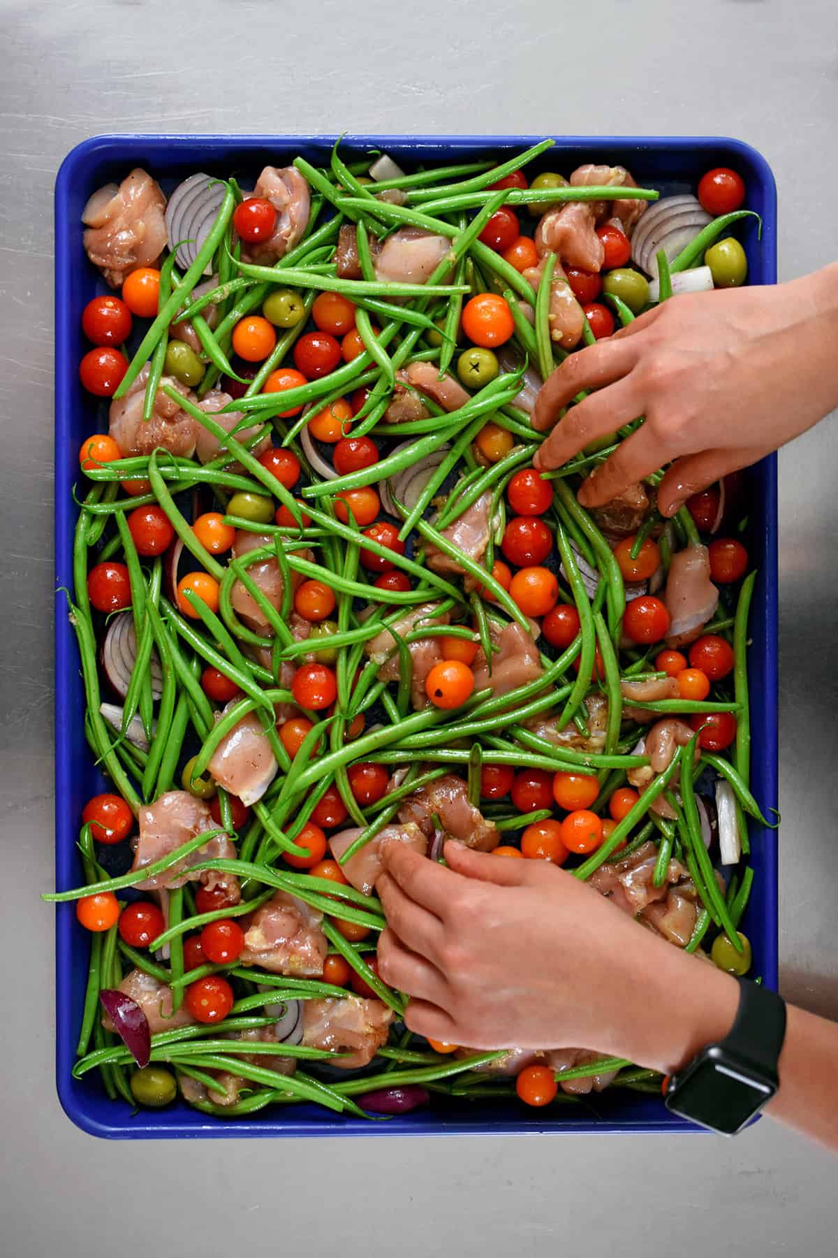 An overhead shot of two hands arranging the ingredients for sheet pan Italian chicken into a single layer on a blue rimmed. baking sheet.