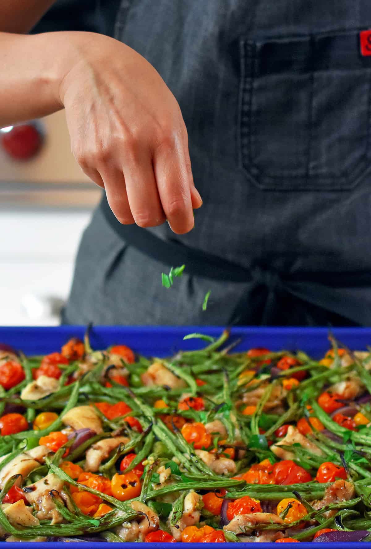 A hand is adding minced fresh herbs to a sheet pan Italian chicken dinner right out of the oven.