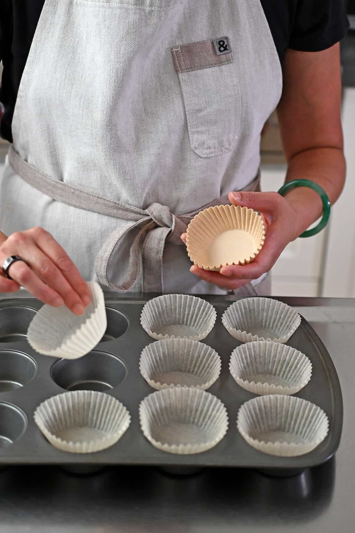 A person in a beige apron is placing parchment liners into a muffin tin.