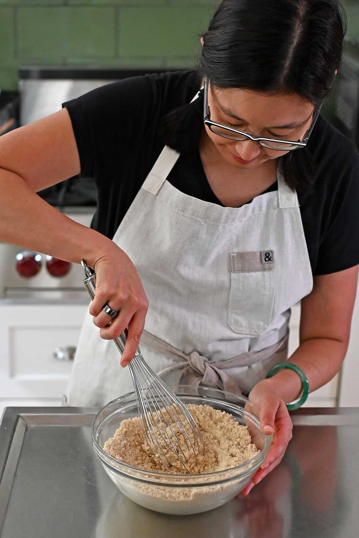 An Asian woman in glasses is mixing the dry ingredients to make paleo apple muffins in a clear glass mixing bowl with a metal whisk.