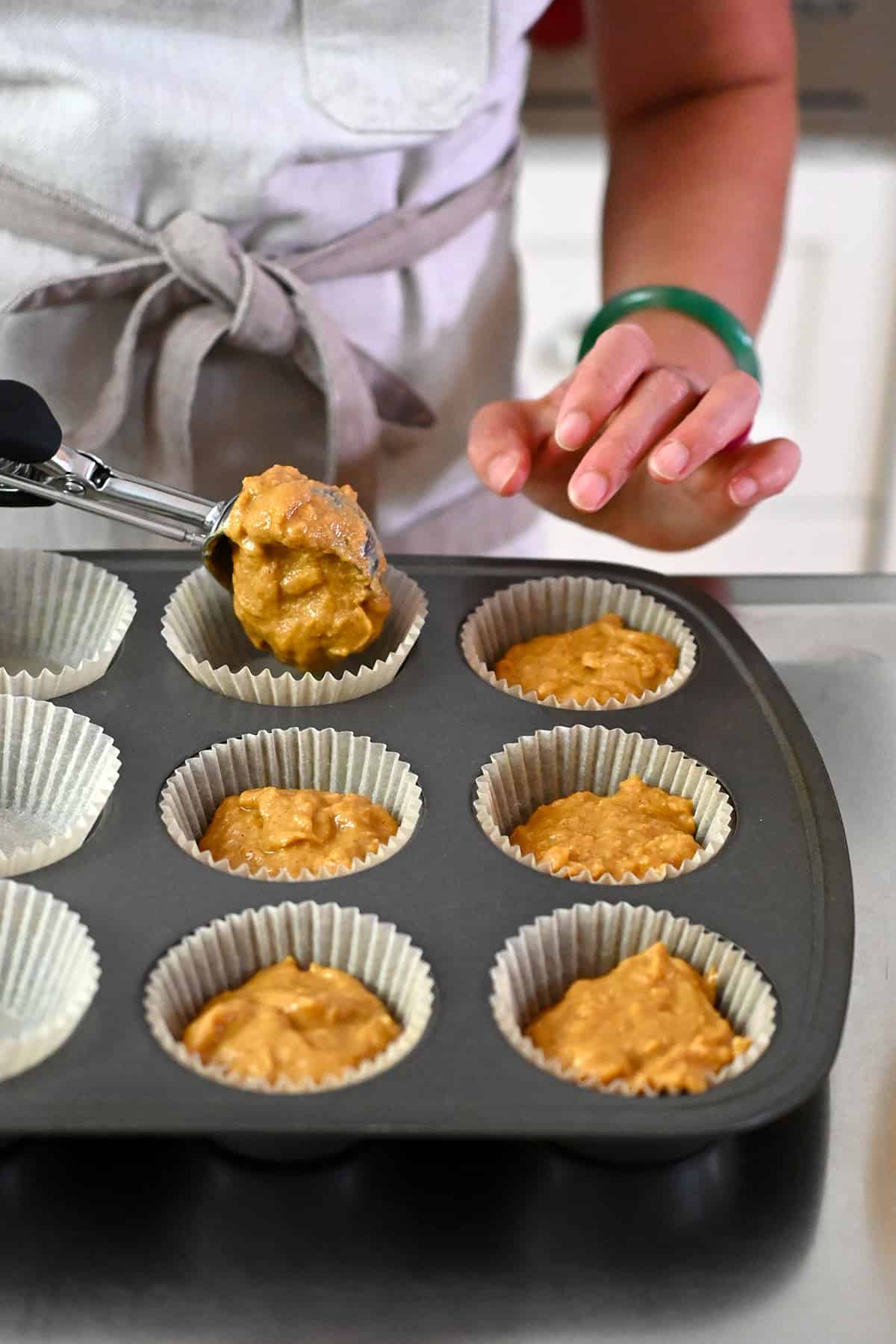 Adding the apple muffin batter with an ice cream scoop to parchment muffin liners in a muffin tin.