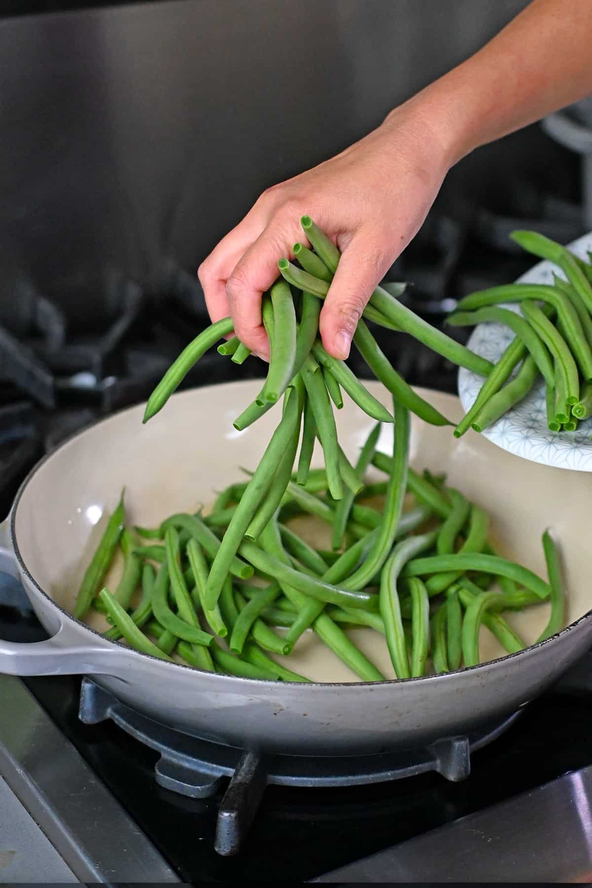Adding trimmed green beans to an enamel cast iron skillet with hot oil.