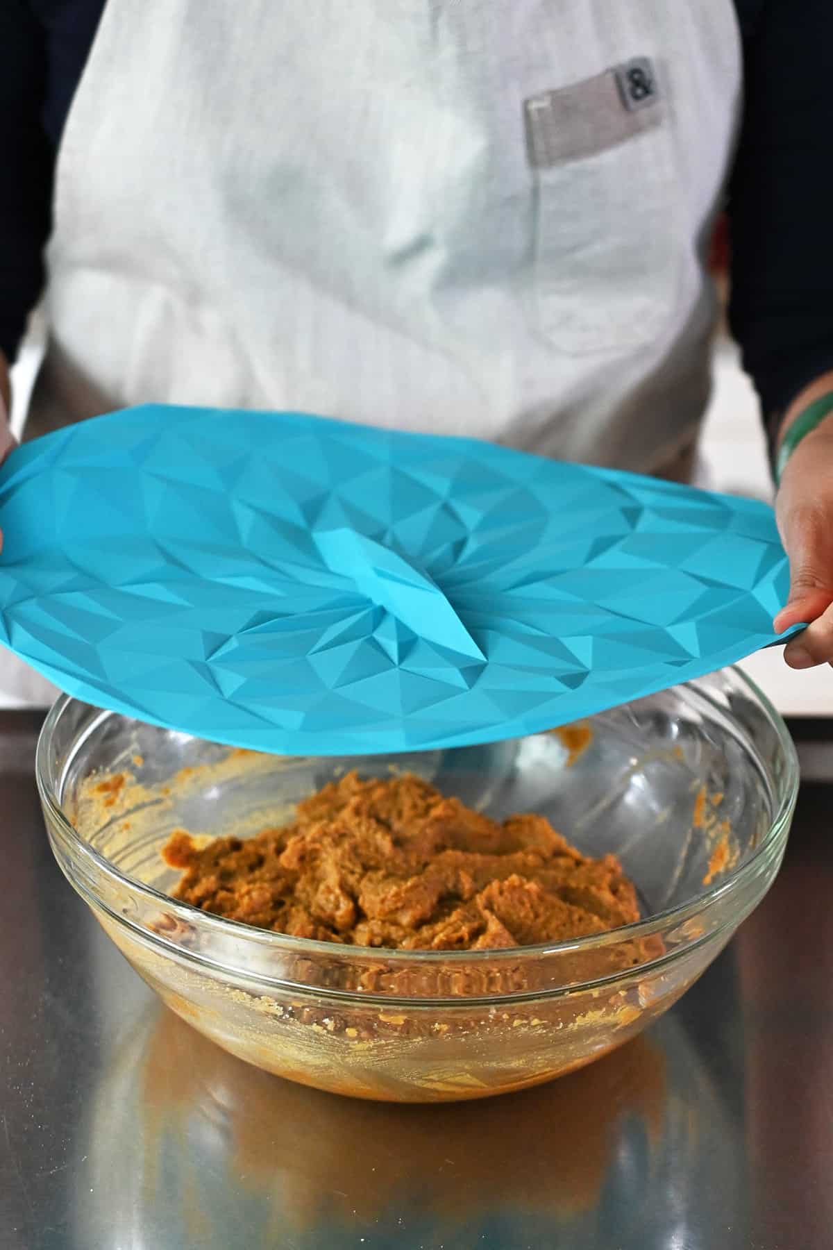 A person is placing a blue silicone lid on top of a bowl filled with uncooked paleo pumpkin cookie dough before chilling it in the fridge.