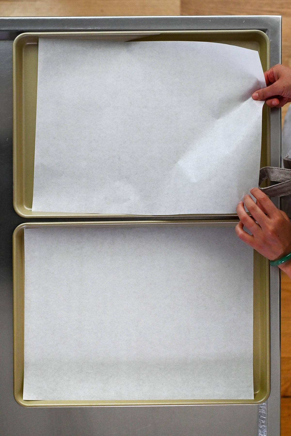 An overhead shot of someone placing parchment paper into two gold colored rimmed baking sheets.