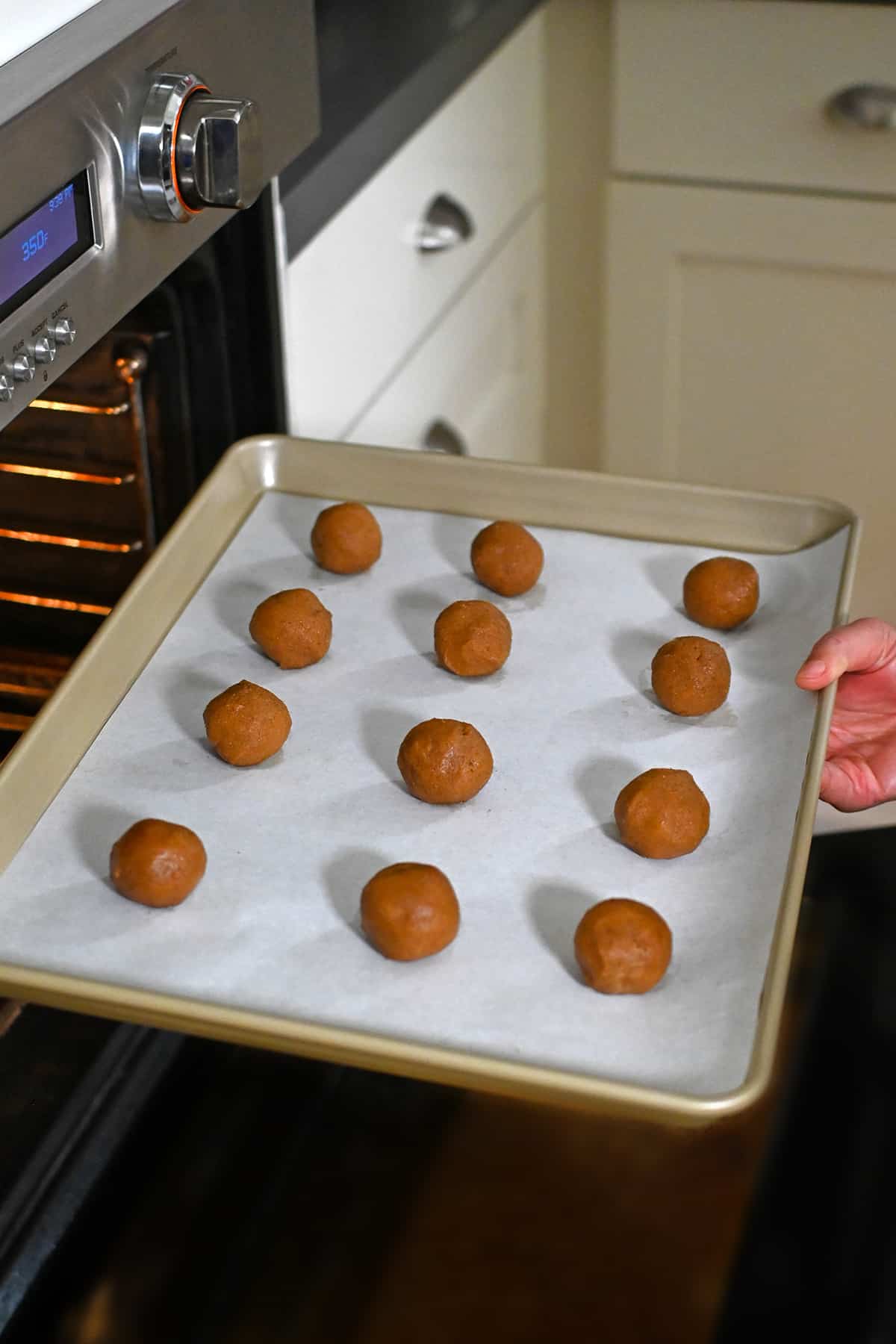 A parchment lined rimmed baking sheet is topped with twelve paleo pumpkin cookie dough balls is being placed into an open oven.