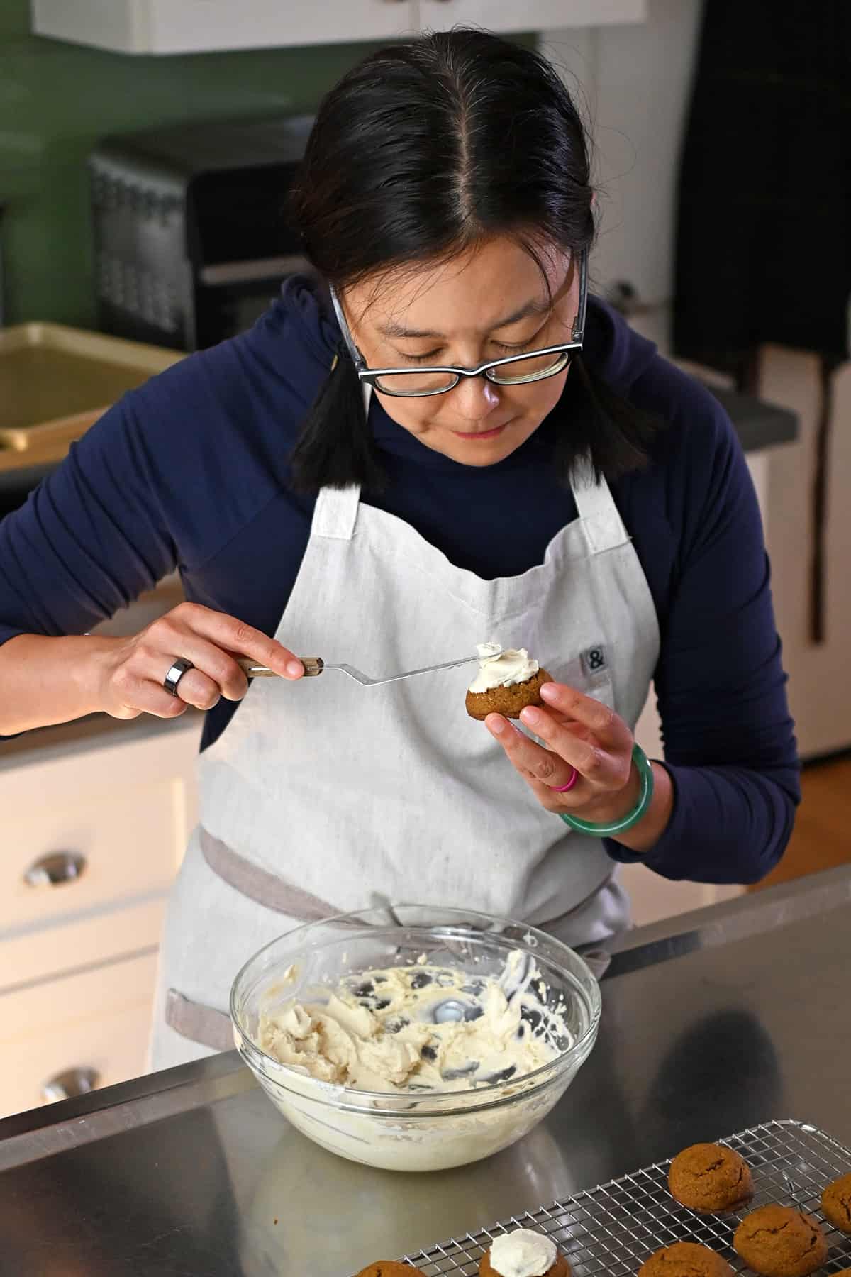 An Asian woman in glasses is using a small offset spatula to add dairy-free cream cheese frosting onto paleo pumpkin cookies.