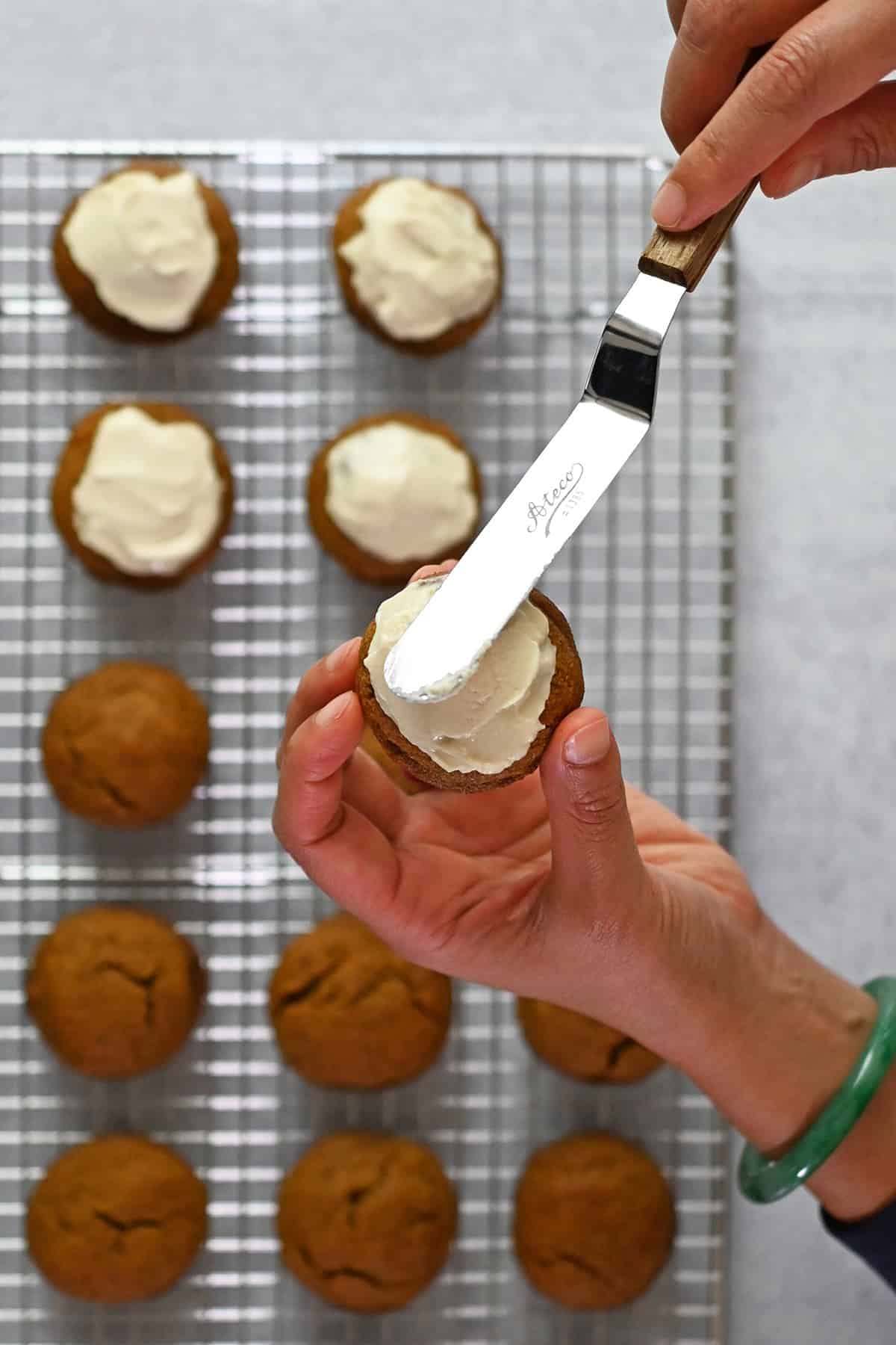 A hand is holding a paleo pumpkin cookie and spreading dairy-free cream cheese frosting on top with a small offset spatula.