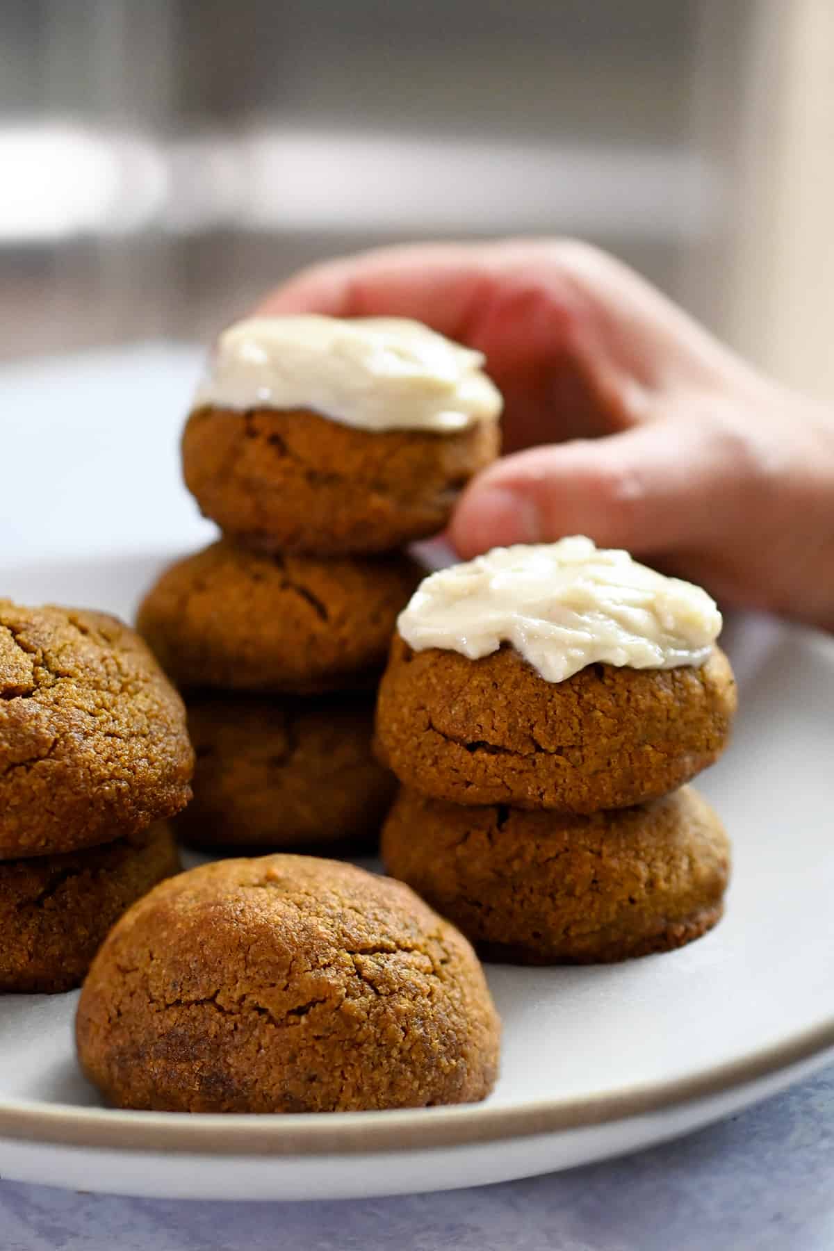 A hand is grabbing a dairy-free cream-cheese frosted pumpkin cookie from a plate filled with cookies.
