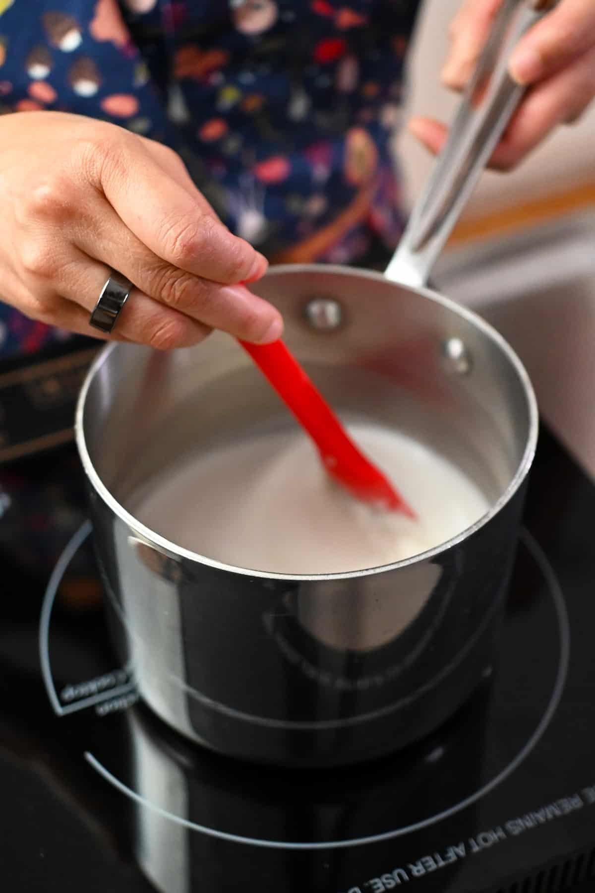 Stirring coconut milk simmering in a small saucepan with a red spatula.