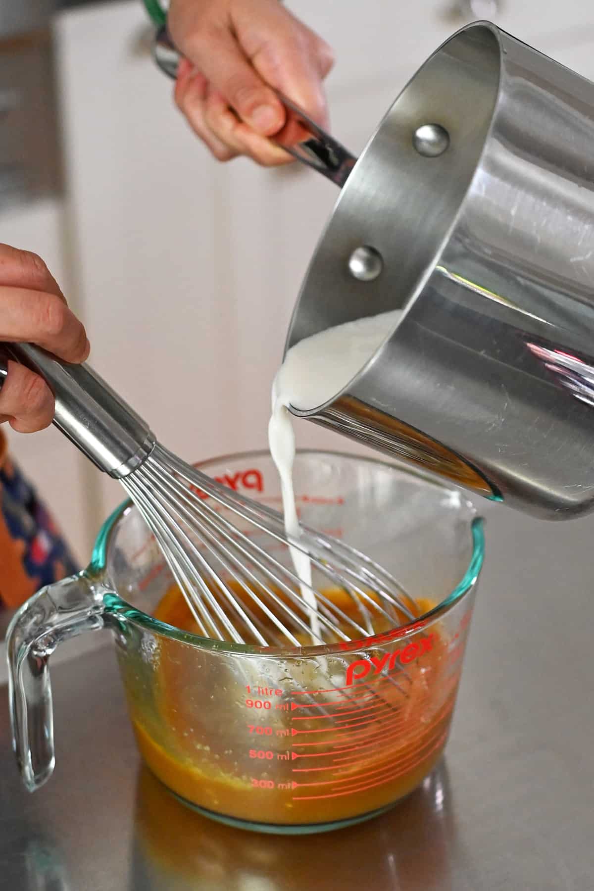 Pouring hot coconut milk into a measuring cup with whisked eggs and maple syrup.